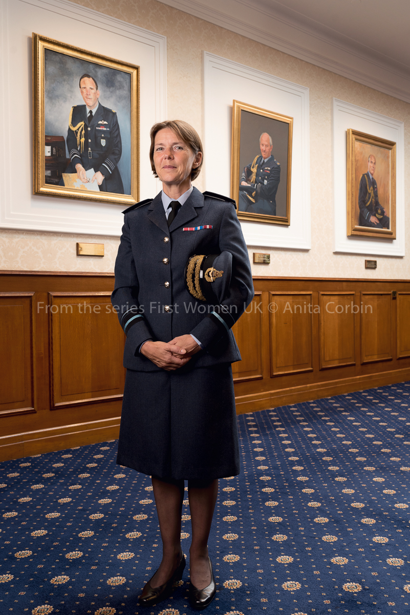 A woman wearing RAF uniform standing in a room with blue carpets and three large portraits hung on the hall behind.