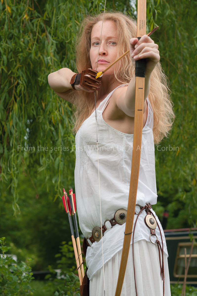 A woman in a white top firing a bow and arrow outdoors.