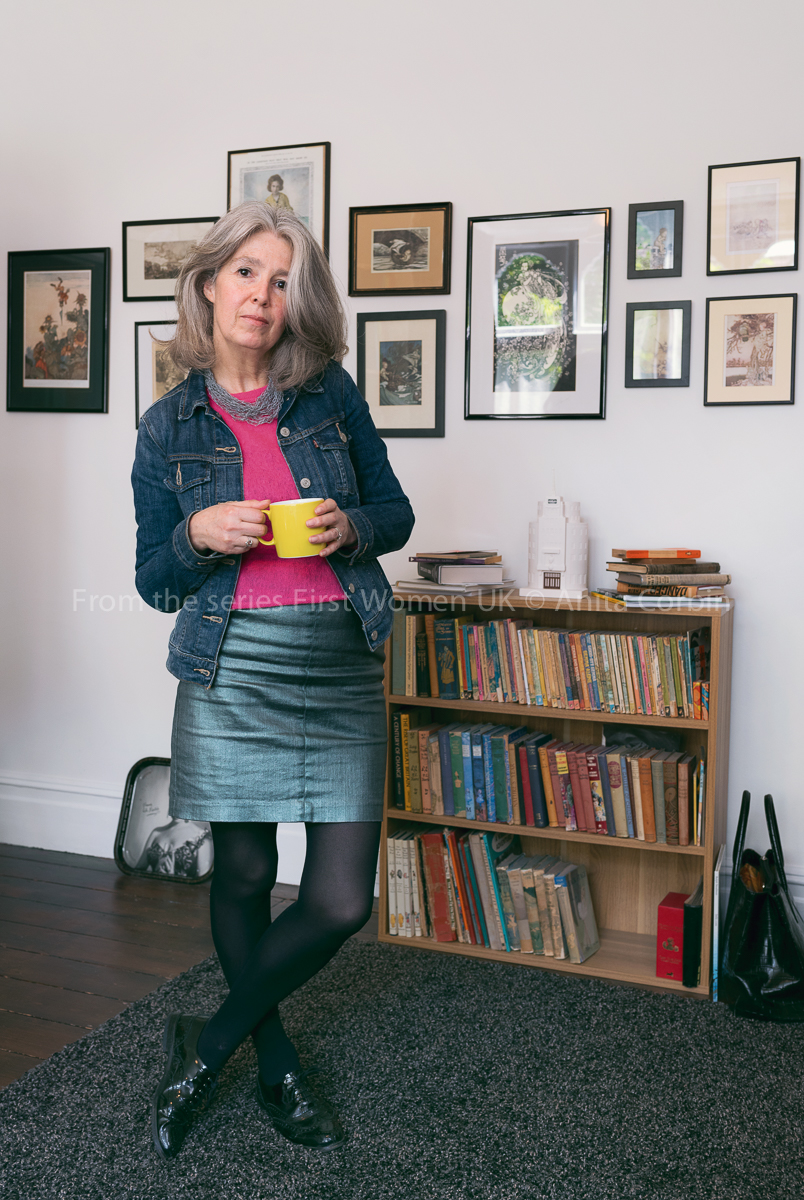 A woman wearing a denim skirt, denim jacket and pink top holding a yellow mug and standing in front of a bookshelf.