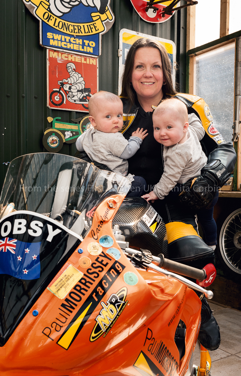 A woman sitting on an orange motorbike with two babies in her arms.
