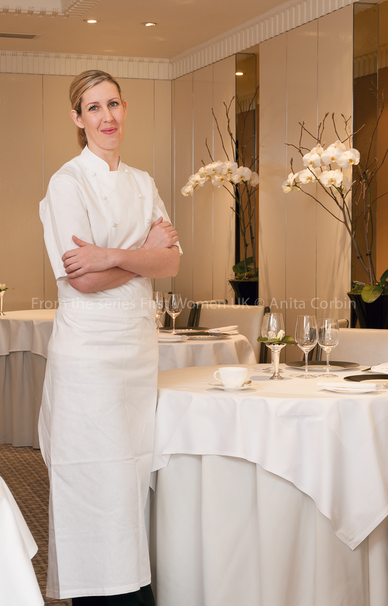 A woman wearing a white chef's outfit with her arms crossed standing next to a small round table with a white tablecloth.