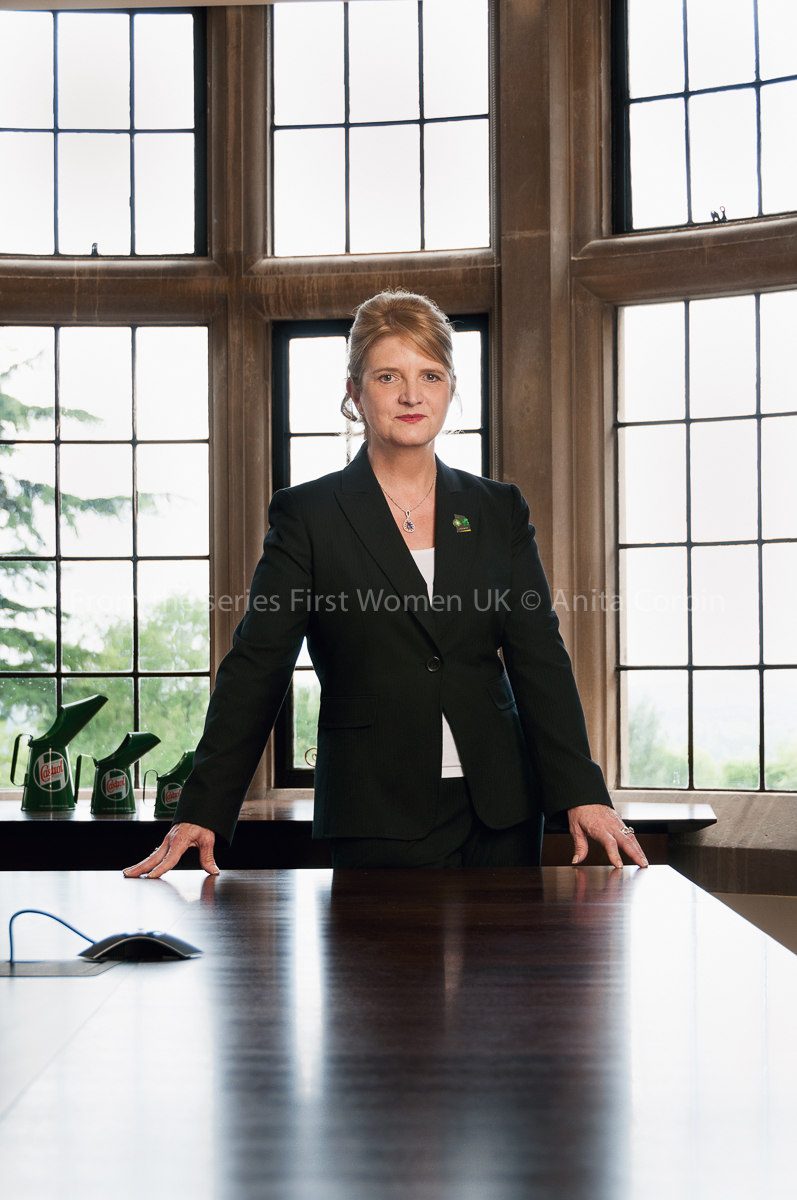 A woman wearing a black suit standing behind a large wooden desk with six window panes behind her.