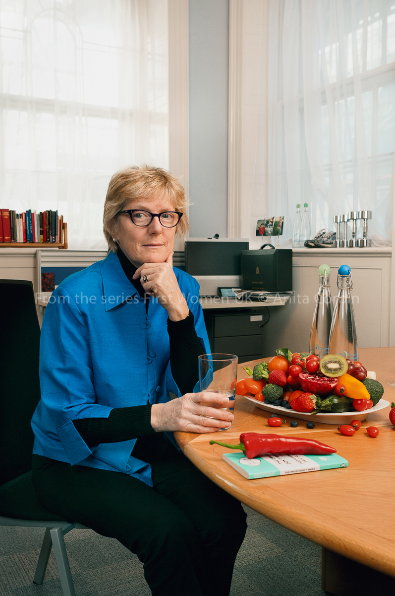 A woman wearing a bright blue shirt, black trousers and glasses sitting at a stable with a large bowl of fruits and vegetables with a glass of water in her hand.