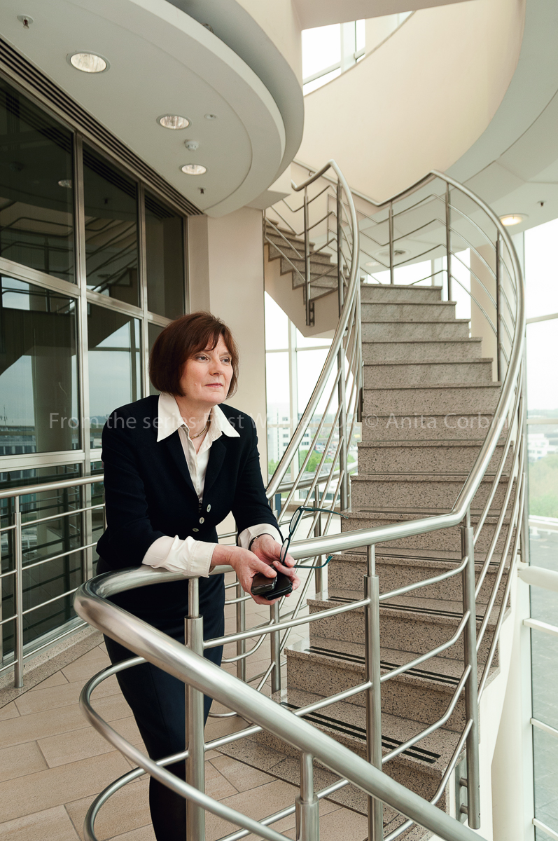 A woman standing with her hands resting on a railing standing at the bottom of a curved staircase in a glass building.