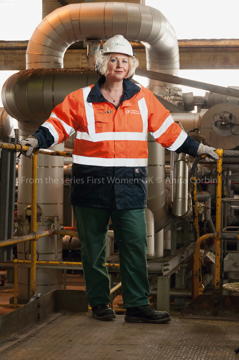 A woman wearing an orange and navy blue waterproof jacket and a white hard hat standing in front of large pipes.