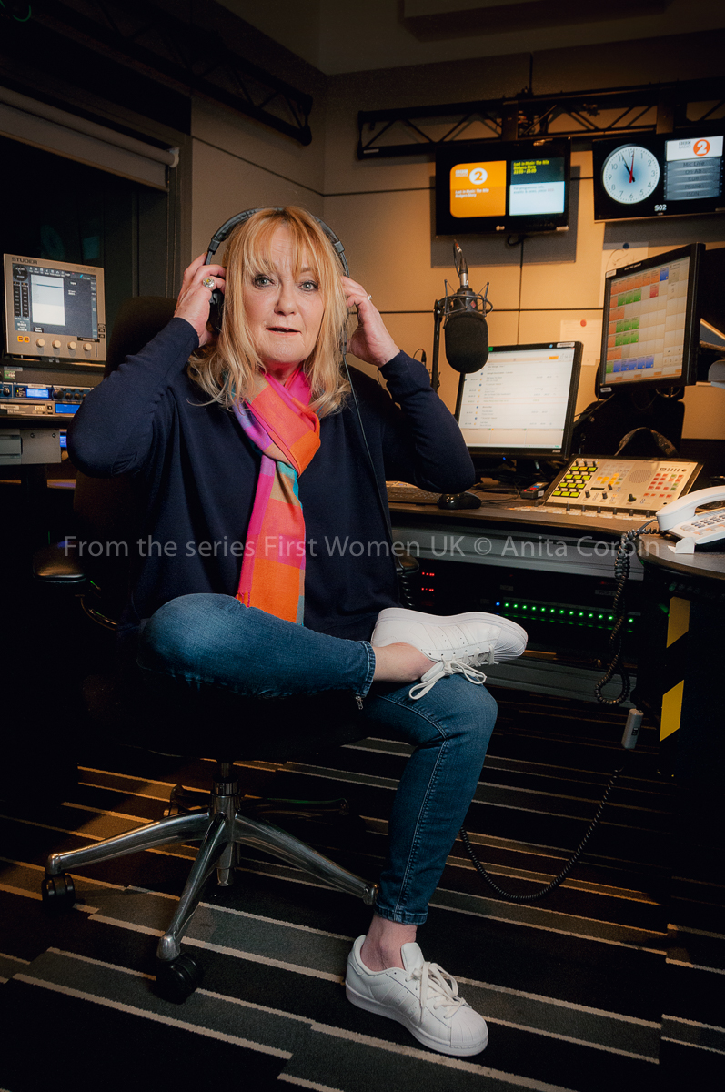 A woman wearing jeans and a pink and orange scarf with one foot on the other knee. She is putting a pair of headphones on her head and sitting in a DJ booth.