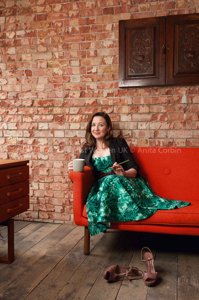 A woman wearing a green dress and black blazer sitting on a red sofa in front of a brick wall. She is holding a grey mug in one hand. A pair of high heeled shoes are scattered on the floor in front.