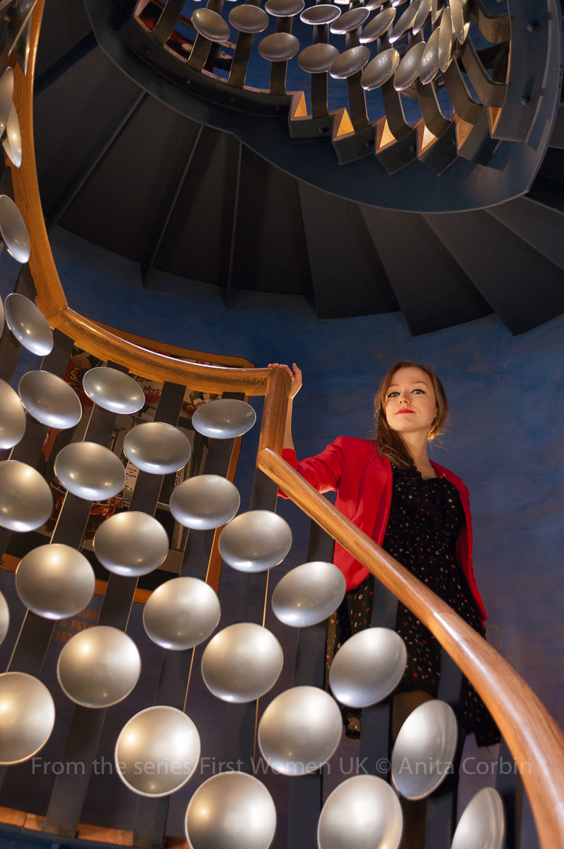 A woman wearing a red blazer standing holding the railing of a curved staircase. The banisters are decorated with silver disks.