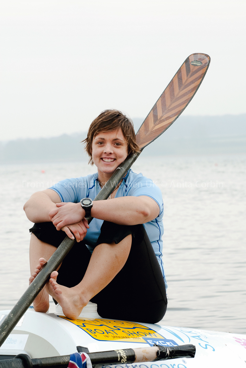 A woman in a light blue top sitting on a rowing boat with an oar over her left shoulder.