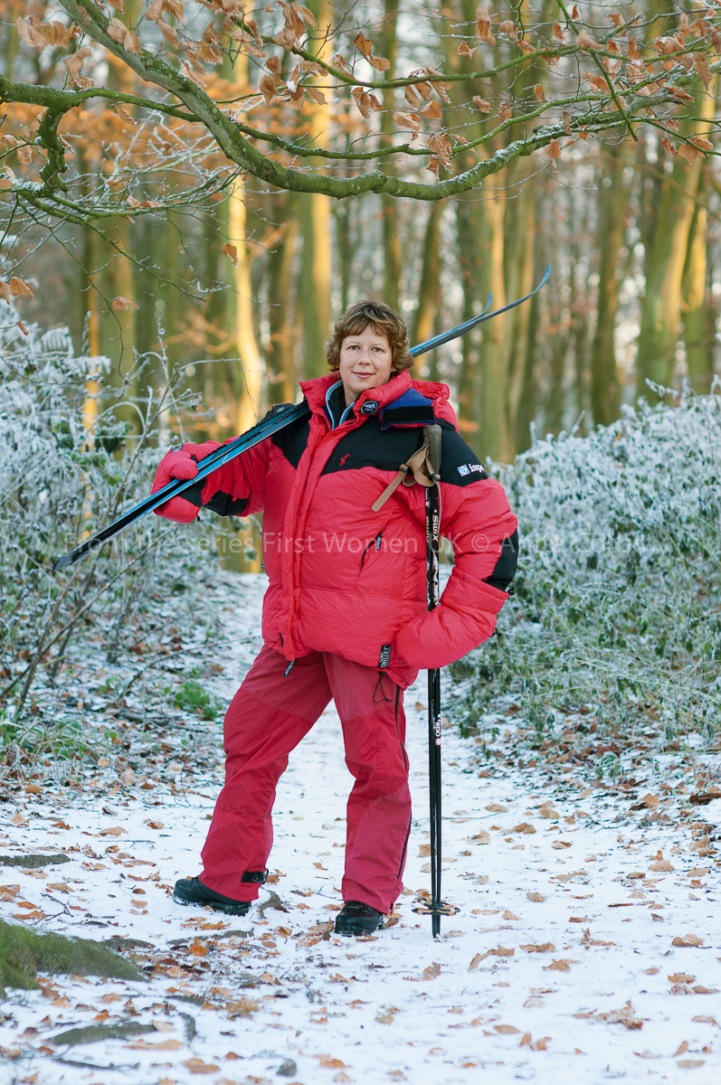 Woman wearing a large red winter coat and matching trousers with skis over her right shoulder. A light dusting of snow on the ground and bushes in the background..